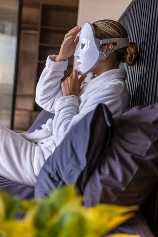 Woman adjusting led facial mask relaxing in white bathrobe on bed for skincare home spa