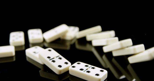 White domino pieces are scattered on a reflective black surface, with a shallow depth of field focusing on the central piece. Dominoes are often used in games of strategy and skill, and this setup suggests a game either in progress or concluded.