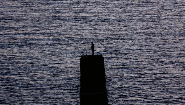 Solitary angler standing on concrete pillar at dusk fishing silhouette over twilight sea