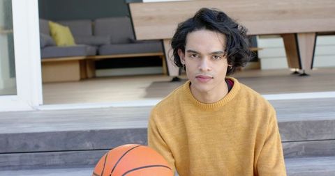 Young man in mustard sweater holding basketball on wooden steps outside modern home