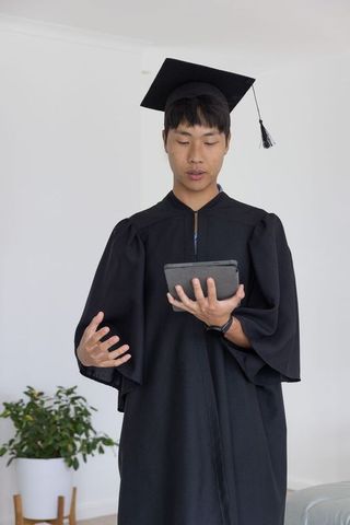 Asian graduate in mortarboard and gown holding tablet delivering virtual graduation speech