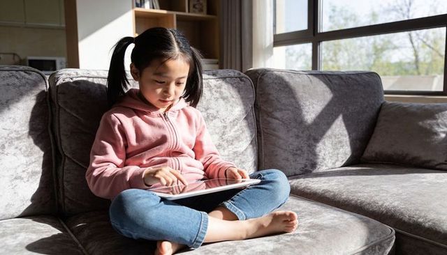 Young girl sitting on sofa using tablet in sunlit living room for online learning and play