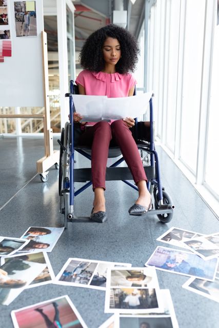 African American graphic designer in a wheelchair reviewing photographs in a modern office. Ideal for themes of diversity, inclusion, professional workspaces, and creative industries. Useful for articles, blogs, and marketing materials focusing on inclusive work environments and creative professions.