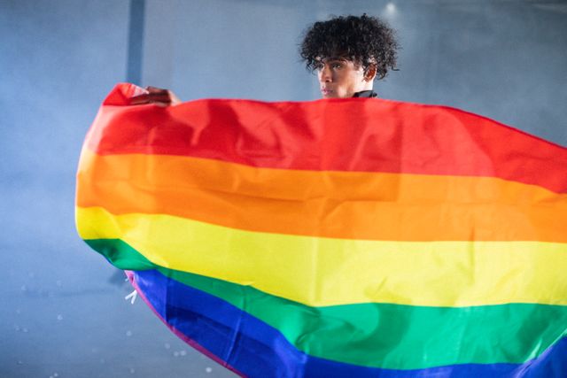 African American transgender man holding a vibrant LGBT flag in an empty parking garage. Ideal for use in campaigns promoting diversity, gender expression, and LGBTQ+ rights. Suitable for articles, blogs, and social media posts about inclusivity and empowerment.