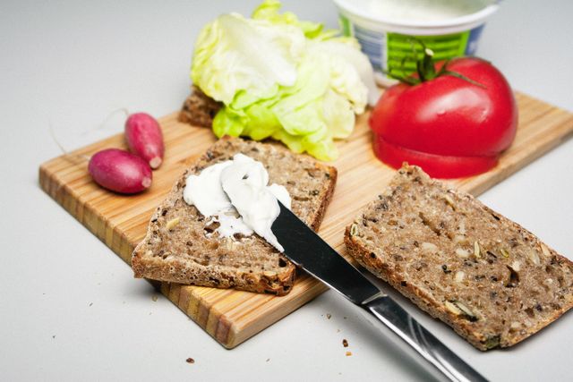 Whole grain bread slices being spread with cream cheese on a wooden cutting board, accompanied by fresh vegetables like radishes, lettuce, and a tomato. Ideal for illustrating healthy eating habits, vegetarian diet, homemade meal preparation, or food-related articles.