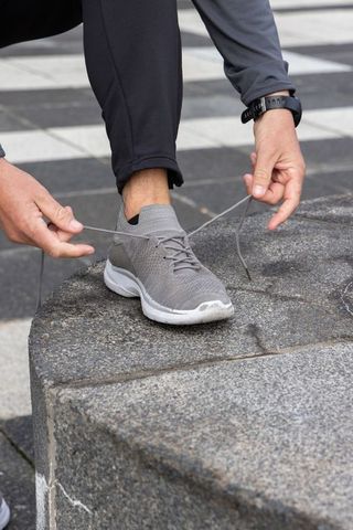 Man tying gray knit running shoe on urban stone block wearing fitness watch in outdoor plaza