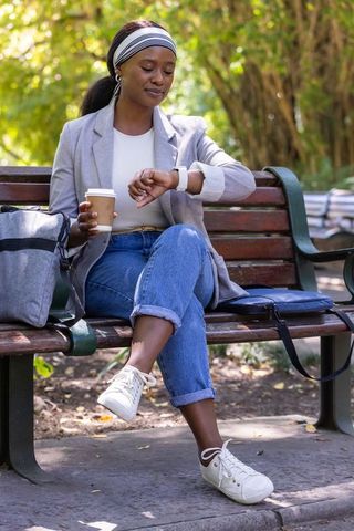Checking smartwatch on park bench while holding coffee, casual blazer and jeans