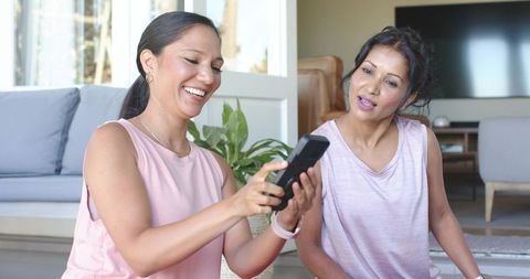 African american and indian women laughing and sharing remote on living room rug