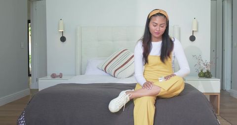 Asian woman in yellow jumpsuit sitting on bed holding remote in modern minimalist bedroom