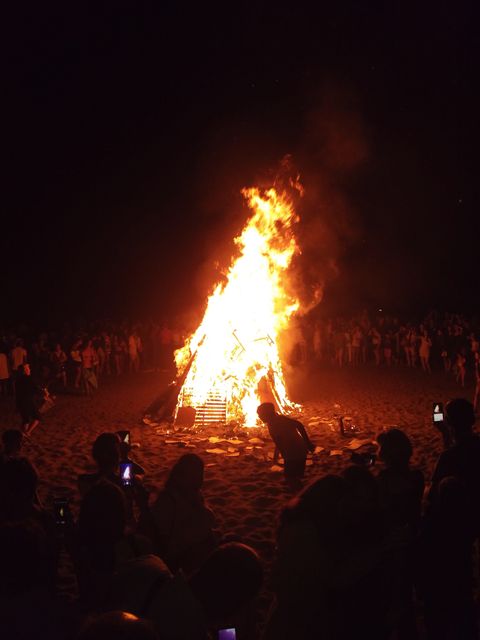 People gathered at night around a large bonfire on a beach. Tall flames rise high, illuminating the dark surroundings and casting a warm glow on the crowd. Perfect for depicting community events, celebrations, or festivals involving beach gatherings and bonfires.