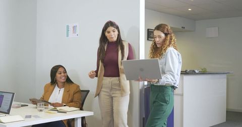 Multicultural female team collaborating with laptop and planner in modern small office