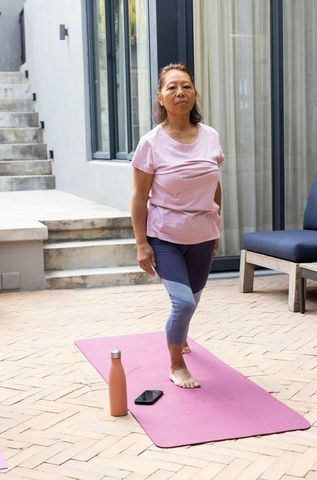 Senior asian woman practicing balance on pink yoga mat outdoors on patio in casual activewear