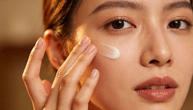 Asian woman applying moisturizer to cheek close-up, glowing skin skincare routine, beauty