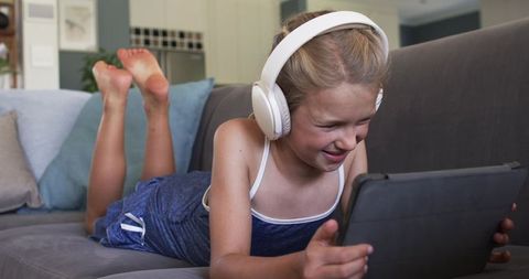 Smiling young girl lying on sofa wearing headphones and using tablet for learning and play