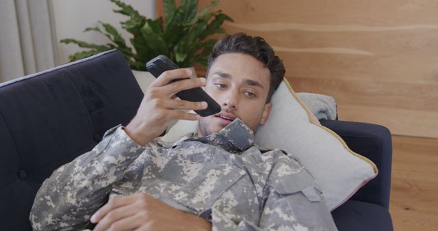 Young army soldier lying on sofa at home, taking break, using smartphone for communication or entertainment. Perfect for content related to military lifestyle, relaxation, veterans, downtime, and modern technology usage.