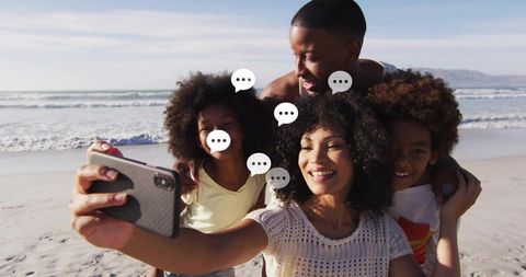 Smiling family taking beach selfie using smartphone with chat bubbles on sunny shoreline