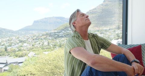 Mature man relaxing on window bench gazing over sunlit valley and suburban panorama
