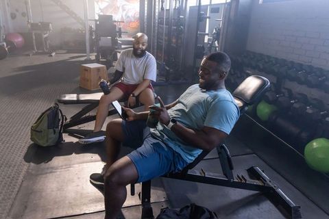 African american men resting on incline bench sharing workout progress on phone