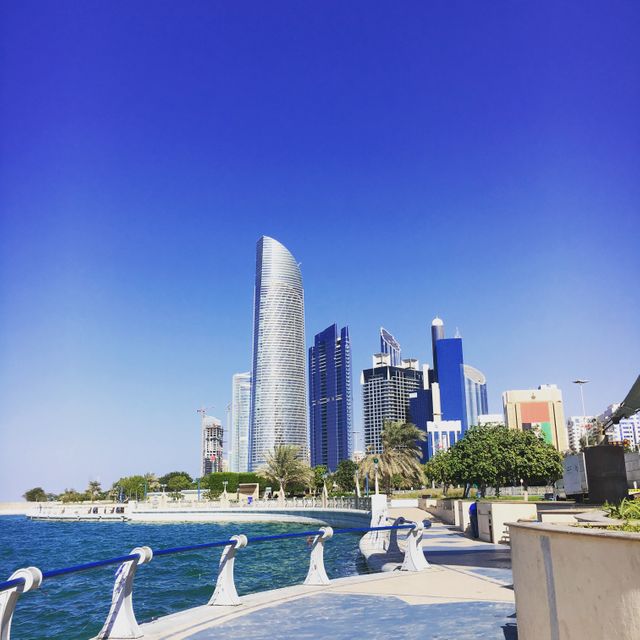 Captures a modern urban skyline with high-rise buildings against a clear blue sky and a waterfront promenade. Suitable for use in travel magazines, tourism promotion, real estate advertisements, business presentations, and blogs focused on contemporary urban living.