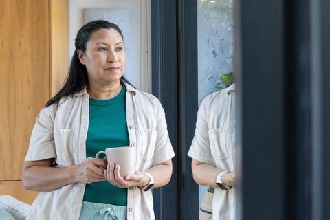 Mature african american woman standing by glass doors holding ribbed mug, contemplative morning