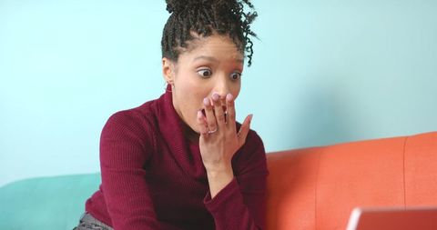 African american woman reacting with surprise, covering mouth while looking at laptop
