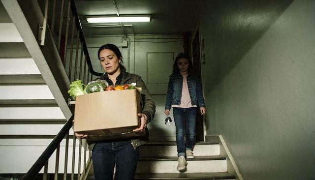 Mother carrying grocery box down dim apartment stairwell with daughter following