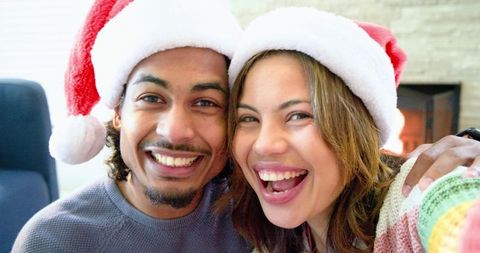 Smiling couple wearing Santa hats and cozy sweaters by fireplace enjoying holiday cheer
