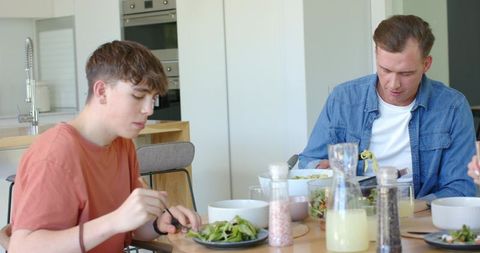 Father and teen son eating healthy salad at modern kitchen table, bright casual meal