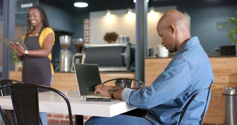 Freelancer working on laptop in urban cafe with african american barista using tablet