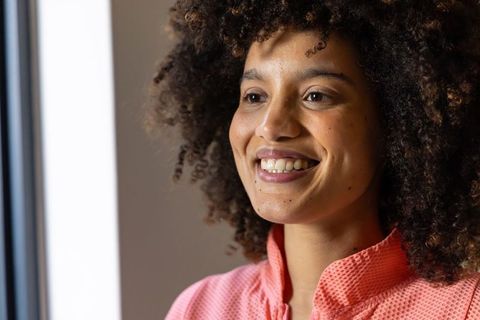 Smiling woman with curly hair looking out window wearing coral collared top