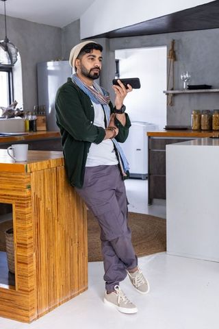 Indian man leaning on bamboo island speaking into smartphone in minimalist modern kitchen