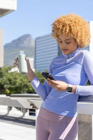 African american woman leaning on rooftop railing checking phone holding water bottle