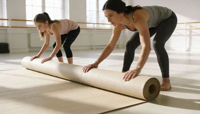 Female instructor and girl rolling yoga mat in sunlit dance studio preparing for class