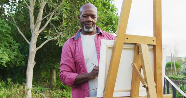 The image shows a mature man engaged in a painting activity in a lush garden setting. He stands before an easel, deeply focused on his work. This image can be used to illustrate topics related to hobbies in later life, art therapy, creative pursuits, or the benefits of spending time in nature.