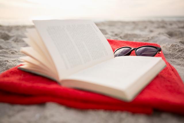 Open book and sunglasses on a red towel at a sandy beach. Ideal for visuals related to summer vacations, relaxation, leisure activities, beach reading, travel ads, and promoting holiday destinations.