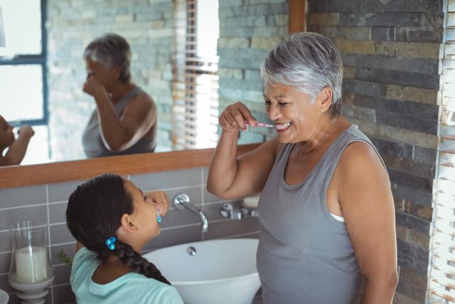 Grandmother and granddaughter brushing teeth together in a modern bathroom. Both are smiling and enjoying the moment, showcasing a strong family bond and daily hygiene routine. Ideal for use in family-oriented content, dental care promotions, and advertisements focusing on intergenerational relationships.