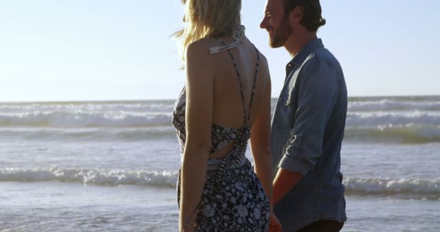 A Caucasian couple enjoys a romantic moment by the sea, with the setting sun casting a warm glow on the horizon. Their peaceful stroll along the beach encapsulates the serenity and connection shared between two people in love.