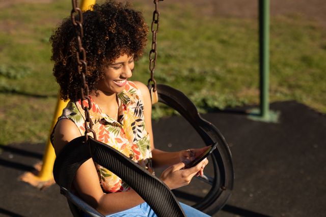 Biracial woman with curly hair sitting on a swing in a playground on a sunny day, using a smartphone and smiling. Ideal for themes related to outdoor activities, leisure, technology use, happiness, and summer lifestyle.
