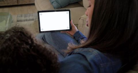 Caregiver holding tablet with child on sofa watching blank screen, cozy home learning