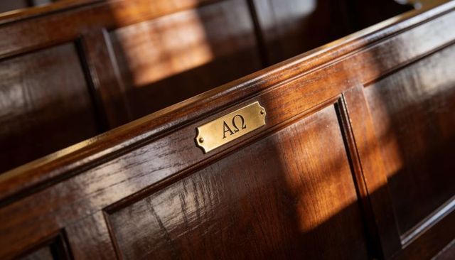Polished wooden church pew with brass alpha omega nameplate and warm sunlight glow