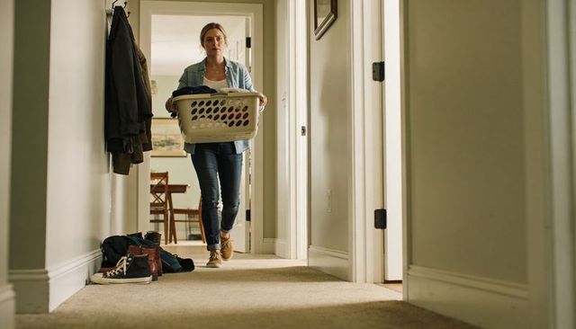 Young woman carrying laundry basket down home hallway in denim shirt and sneakers