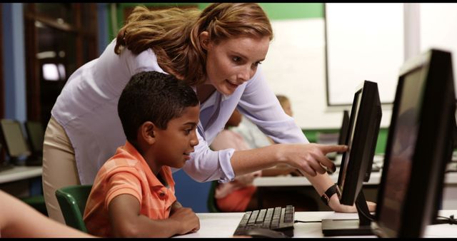 Educator providing one-on-one assistance to a young student during a computer lesson in an elementary school classroom. Suitable for topics on education, mentorship, technology in schools, children's learning, and teaching methods.