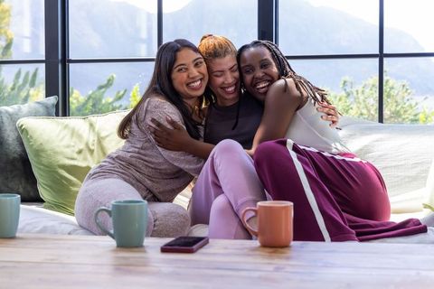 Diverse women friends hugging on sofa in loungewear, cozy morning sunlight and laughter