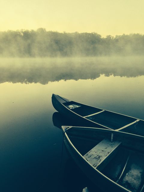 Two canoes are floating on a misty lake at dawn, with the still water reflecting the surrounding landscape. Mist rises from the surface, creating a serene and tranquil atmosphere. Invisible stands of trees can be slightly seen beyond the water’s edge, shrouded in early morning fog. Perfect for use in promoting outdoor adventures, travel destinations, and nature retreats. Ideal for background on websites or newsletters aiming to create a peaceful or inviting impression.