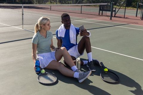 Diverse tennis duo resting on court checking phone hydrating during sunny outdoor break