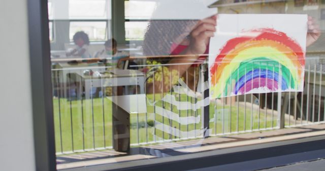 Schoolgirl wearing face mask, glueing a rainbow drawing on a window. Education back to school health safety during Covid19 Coronavirus pandemic.