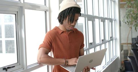African American man standing by large windows typing on laptop in modern coworking space
