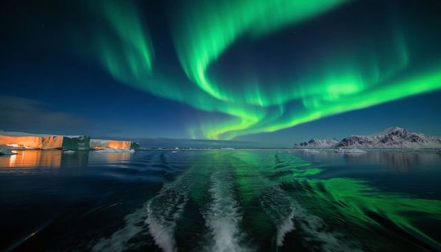 Dancing aurora borealis over arctic waters with boat wake, lit iceberg and snowy peaks