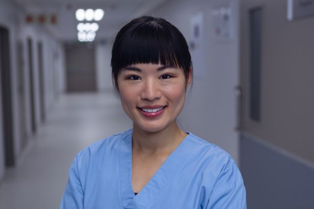 Female surgeon standing confidently in a hospital corridor while smiling. She is wearing blue scrubs, exemplifying readiness and professionalism. Ideal for promoting healthcare, medical services, hospital environments, and professional healthcare staff.