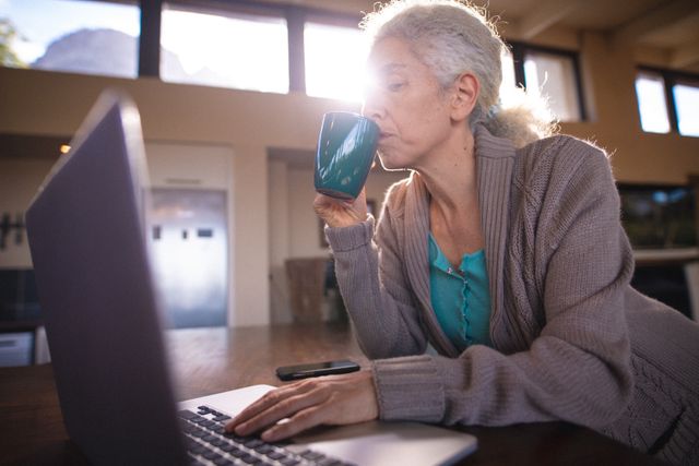 Senior woman enjoying coffee while using a laptop at home. Ideal for illustrating retirement lifestyle, technology use among elderly, and morning routines. Suitable for articles on senior living, home comfort, and independent living.